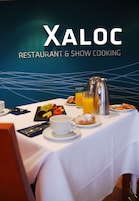 Una mesa de desayuno con diversas opciones de comida y una cafetera, con un fondo que muestra el texto "XALOC RESTAURANT & SHOW COOKING".