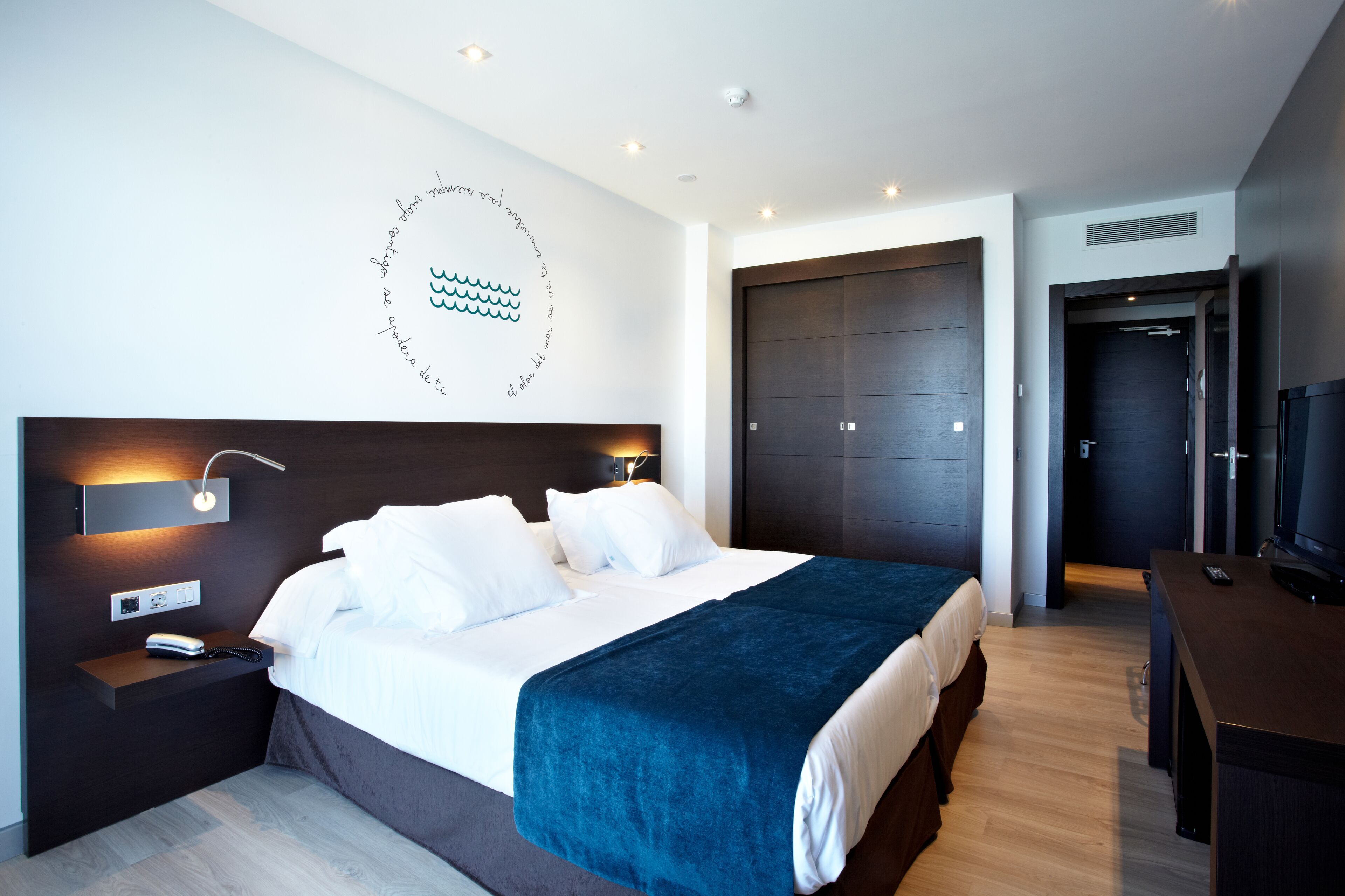 A modern hotel room with a large bed, bedside tables, a TV, and a dark wooden wardrobe.
