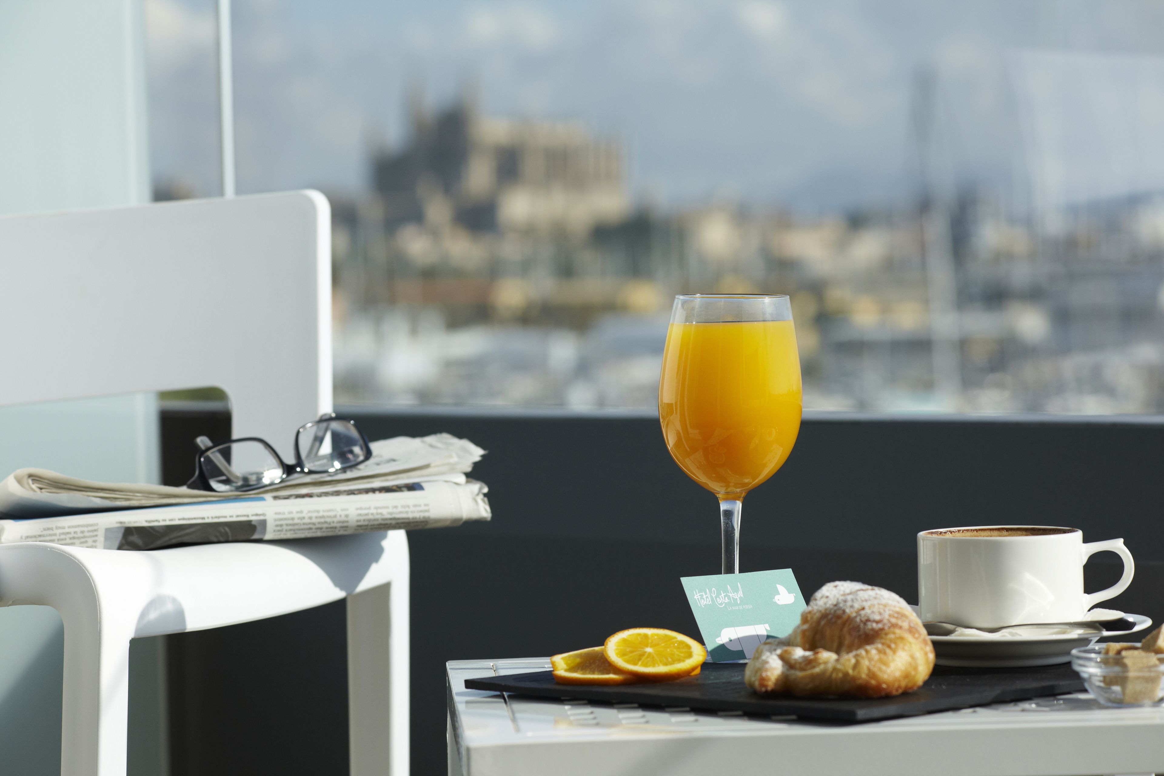 Une table dressée pour le petit-déjeuner, avec un croissant, une tranche d’orange et un verre de jus d’orange, le tout placé devant une fenêtre offrant une vue sur la ville.