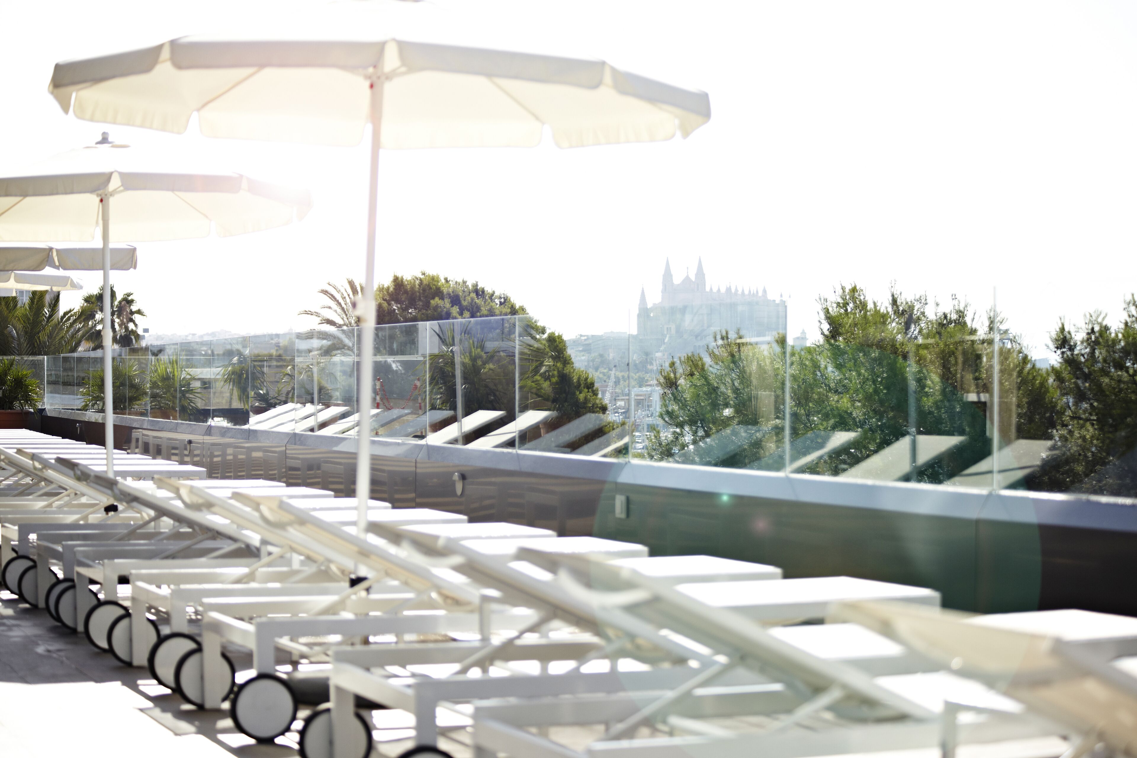 Un espace de détente aménagé sur le toit, avec des chaises longues blanches et des parasols, offrant une vue sur le paysage urbain et un imposant édifice qui ressemble à un château.
