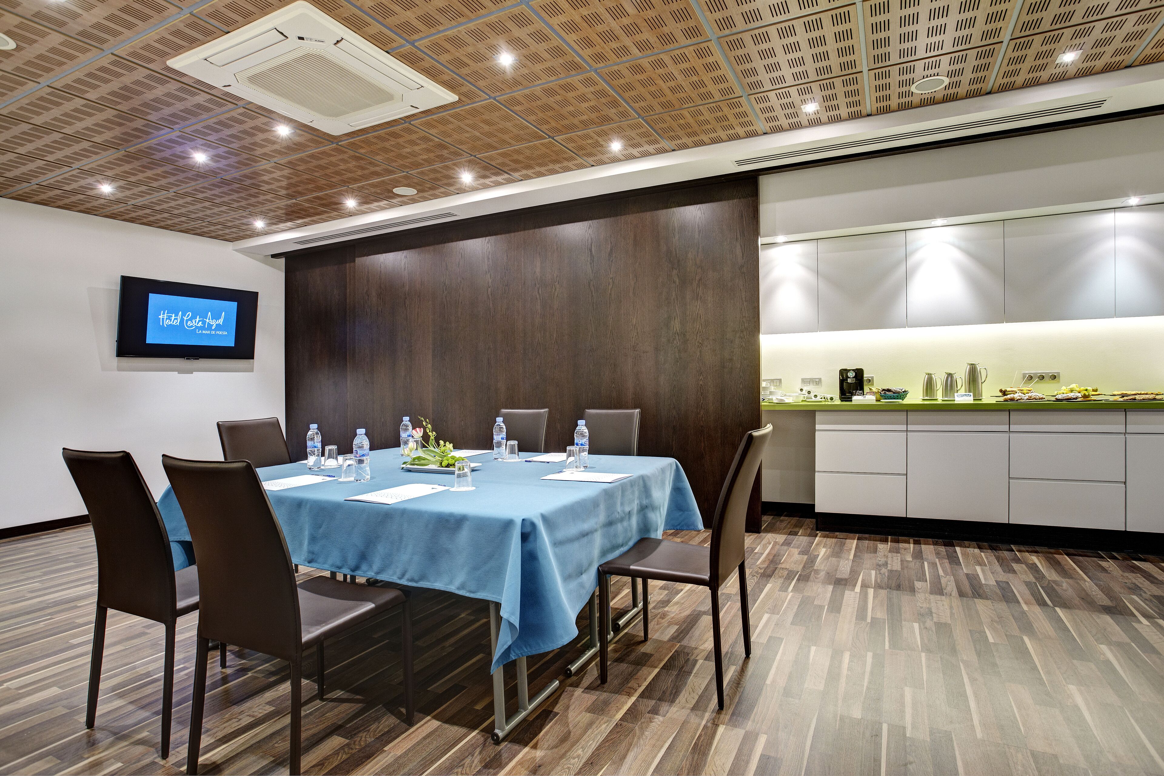 Una sala de conferencias con una mesa azul, sillas marrones, una pantalla de televisión y una zona de cocina.