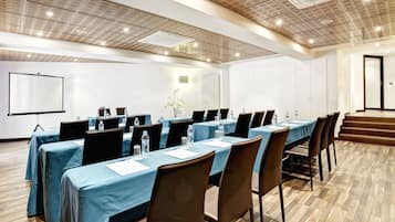 Un salón de conferencias con largas mesas, sillas y una pantalla para proyector.