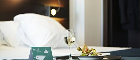 Una cama bien hecha con una almohada blanca, una copa de vino blanco, un plato de comida y una tarjeta sobre un edredón azul.