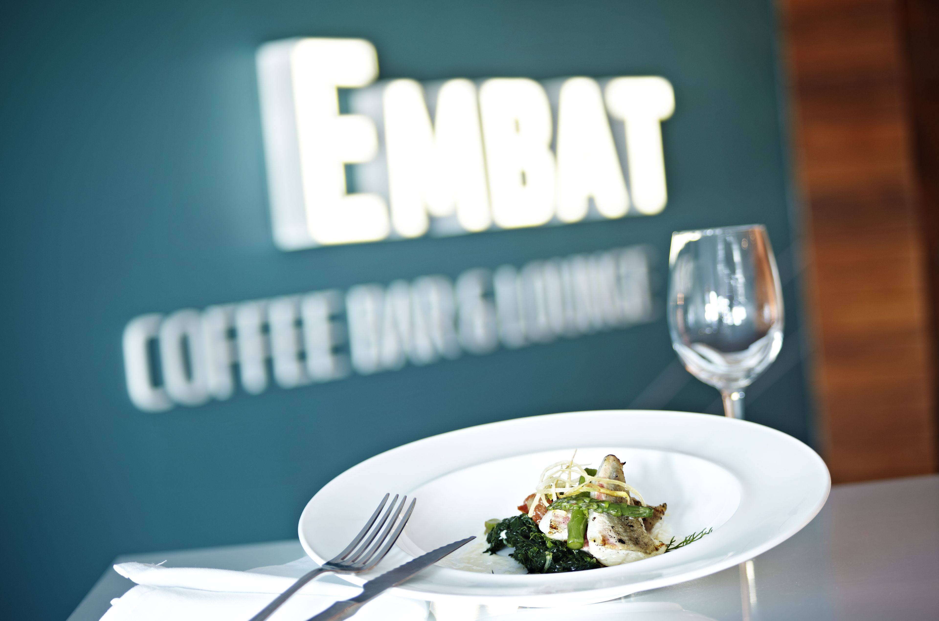 Ein Teller Essen auf einem Tisch mit Gabel und Messer, ein Weinglas und ein Schild im Hintergrund mit der Aufschrift 'EMBAT COFFEE BAR & RESTAURANT'.