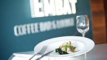 Une assiette de nourriture est posée sur une table, avec une fourchette et un couteau à côté, un verre à vin, et, en arrière-plan, une enseigne indiquant « EMBAT COFFEE BAR & RESTAURANT ».