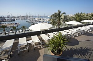 Una terraza en la azotea con sillones blancos, una palmera y vistas a un puerto deportivo con barcos.