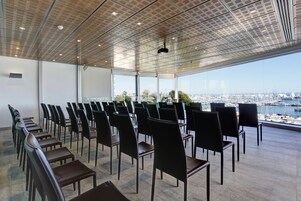 Un salón de conferencias con filas de sillas y mesas, amplias ventanas con vista a un puerto deportivo urbano.