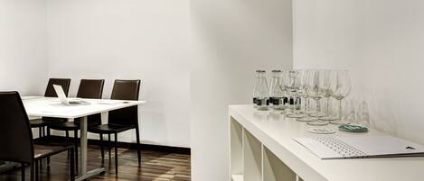 Une salle de conférence avec une table blanche, des chaises noires et un ordinateur portable. On peut voir des verres à vin sur une étagère blanche.