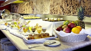 Un bufé con diversos alimentos y bebidas, incluyendo frutas y postres, dispuesto sobre una encimera de mármol con fondo de pared de piedra.