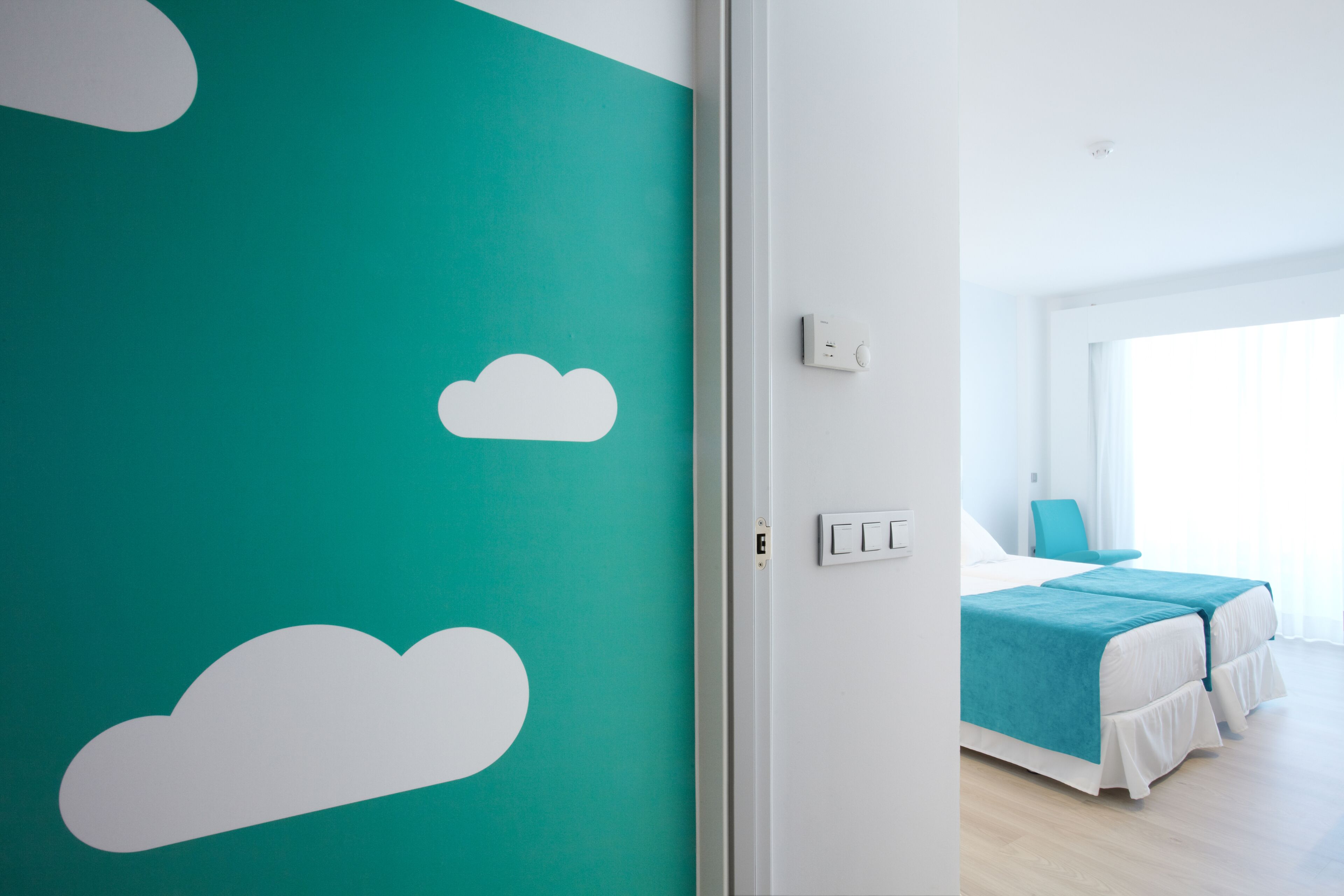 A hotel room with a teal accent wall, a bed with a blue bedspread, and a window with curtains.