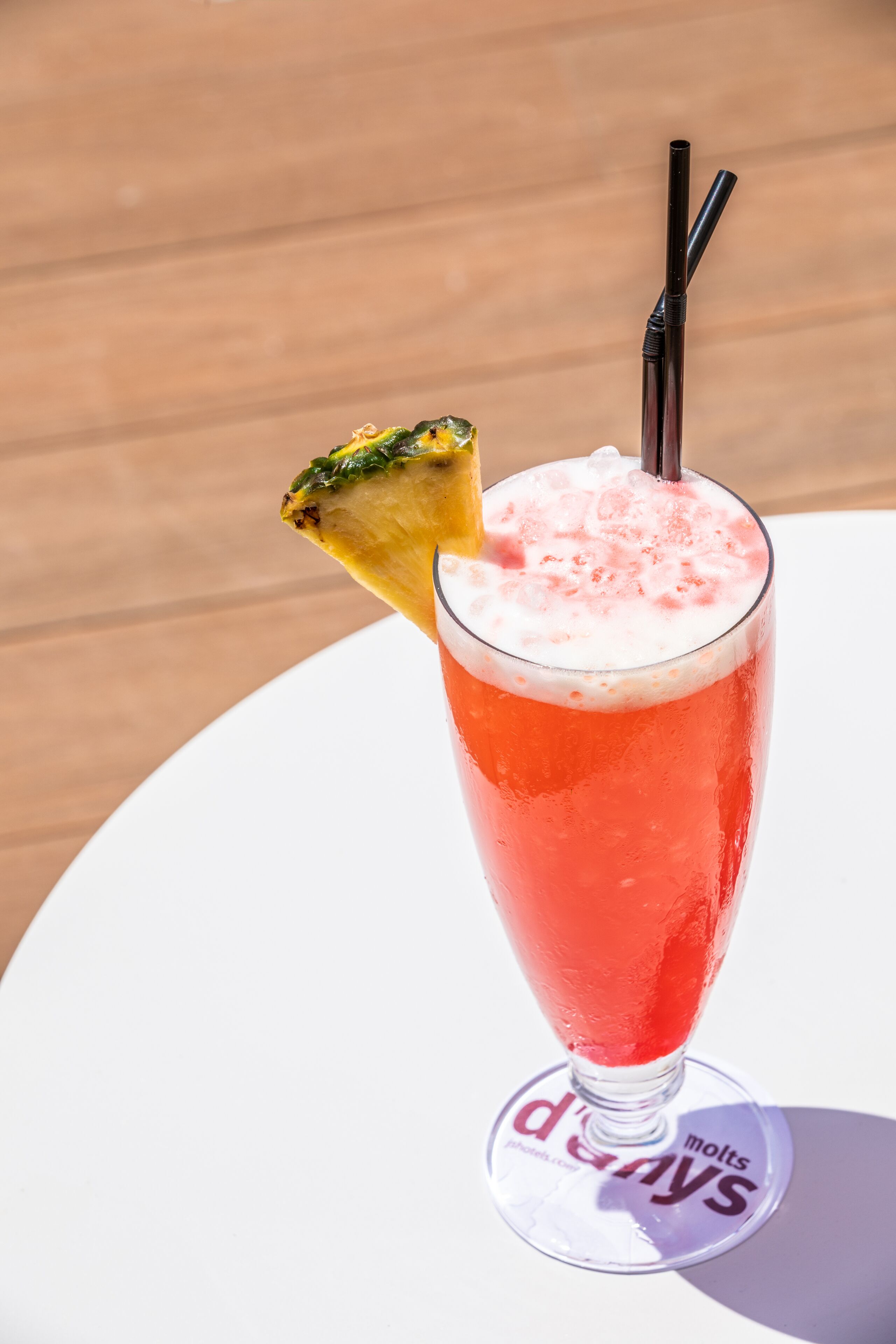 A tall glass with a red beverage, a pineapple slice, and a straw on a white coaster with text.