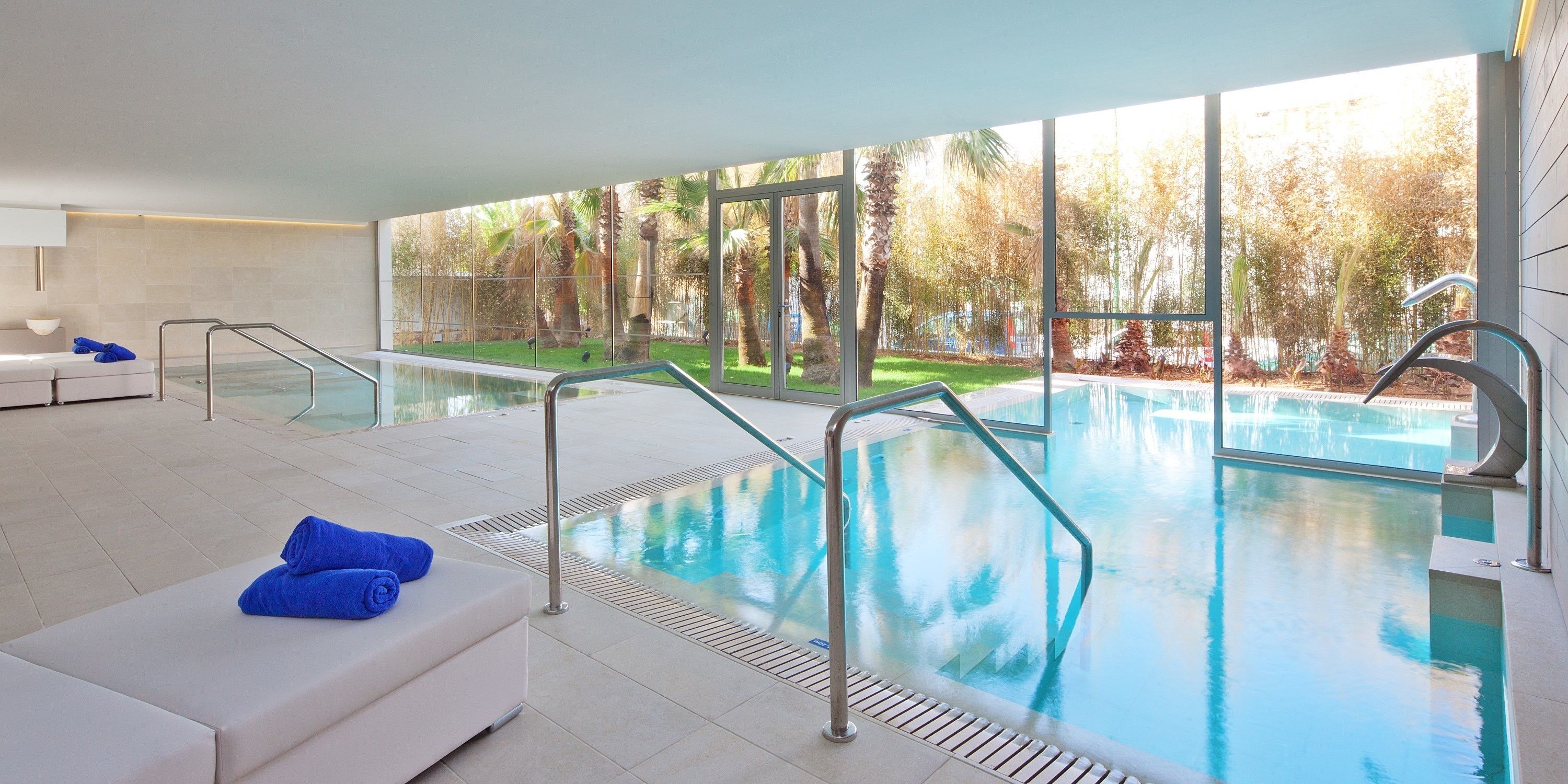 A modern indoor swimming pool with clear glass walls, a white lounge area with blue towels, and a view of a landscaped garden.