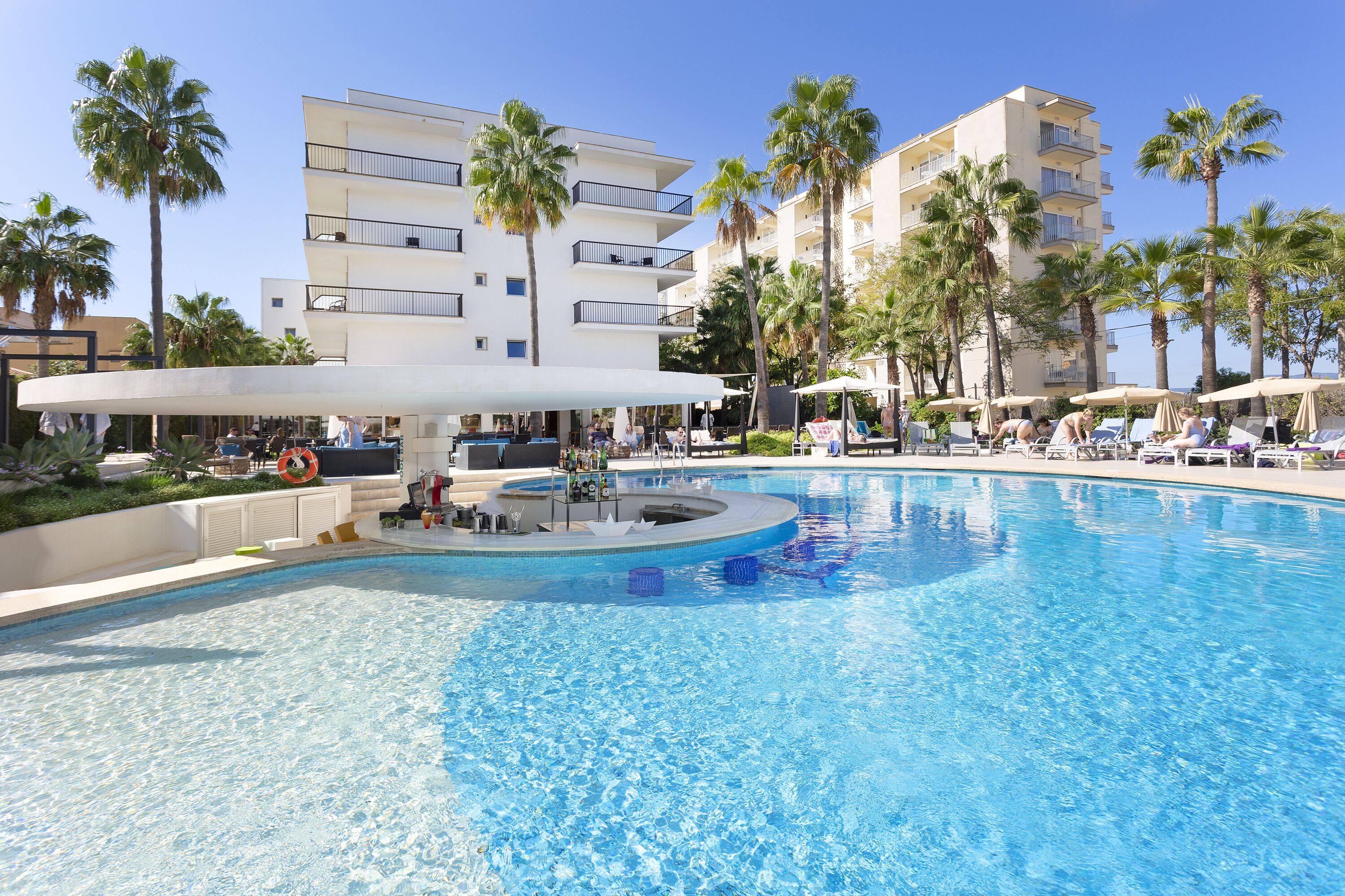 A swimming pool with lounge chairs and umbrellas, surrounded by palm trees and modern buildings.