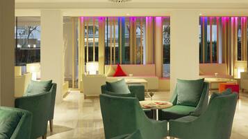 A modern hotel lobby with green upholstered chairs, a sofa with a red pillow, and a table with a glass of wine.