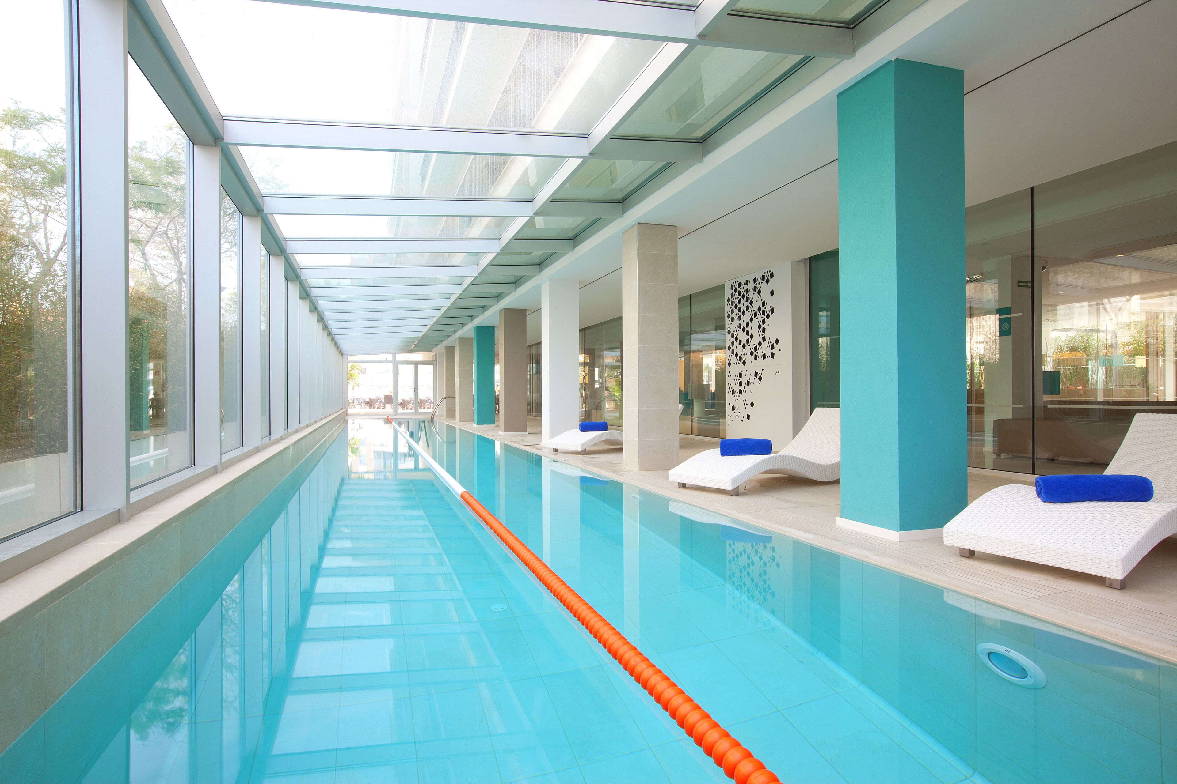 Una lunga piscina interna con pareti di vetro trasparente, acqua azzurra e lettini da relax bianchi con cuscini blu.