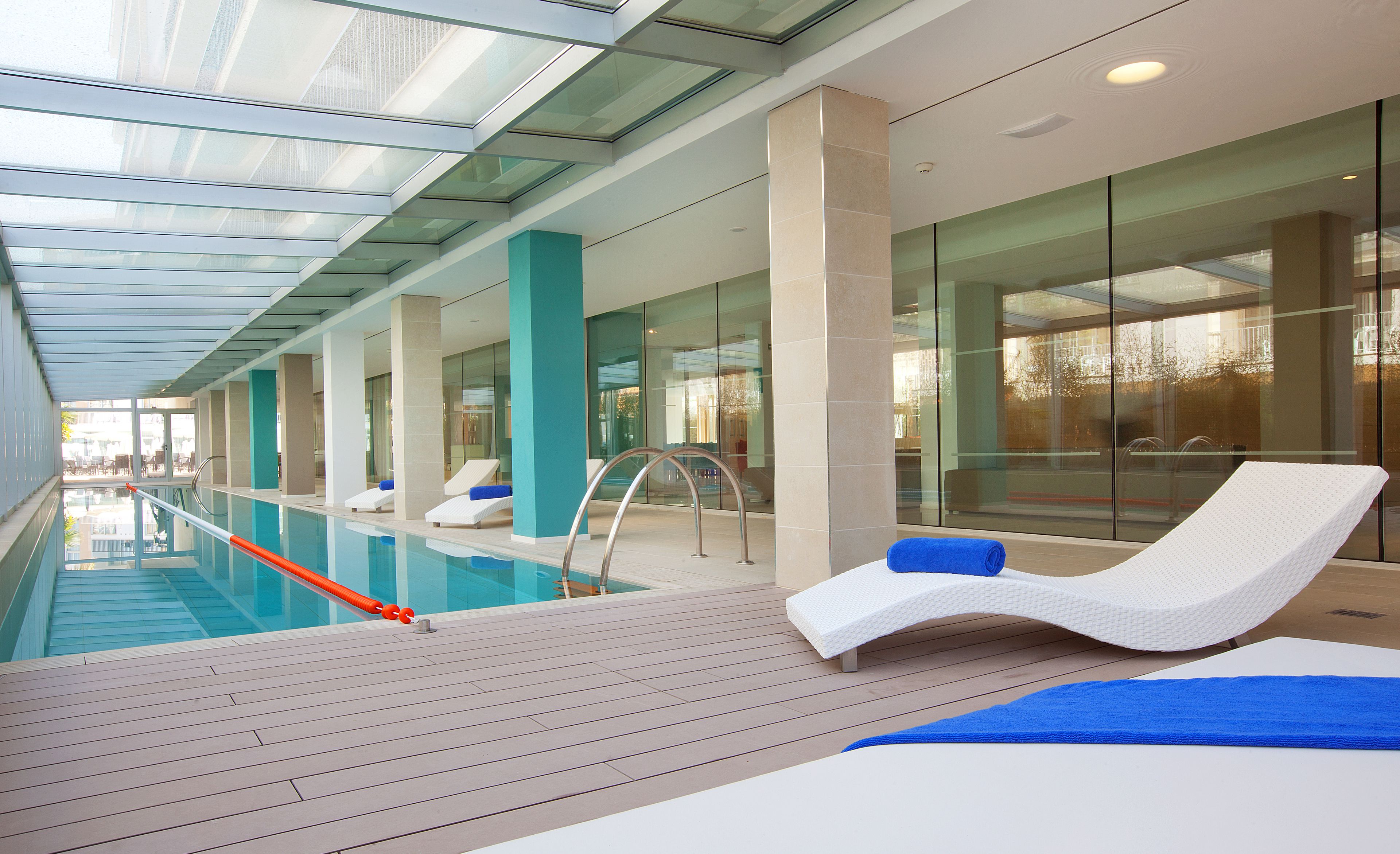 Un área de piscina interior moderna con paredes de vidrio transparente, sillas de descanso y una terraza de madera.