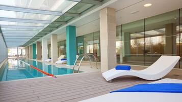 Un área de piscina interior moderna con paredes de vidrio transparente, sillas de descanso y una terraza de madera.