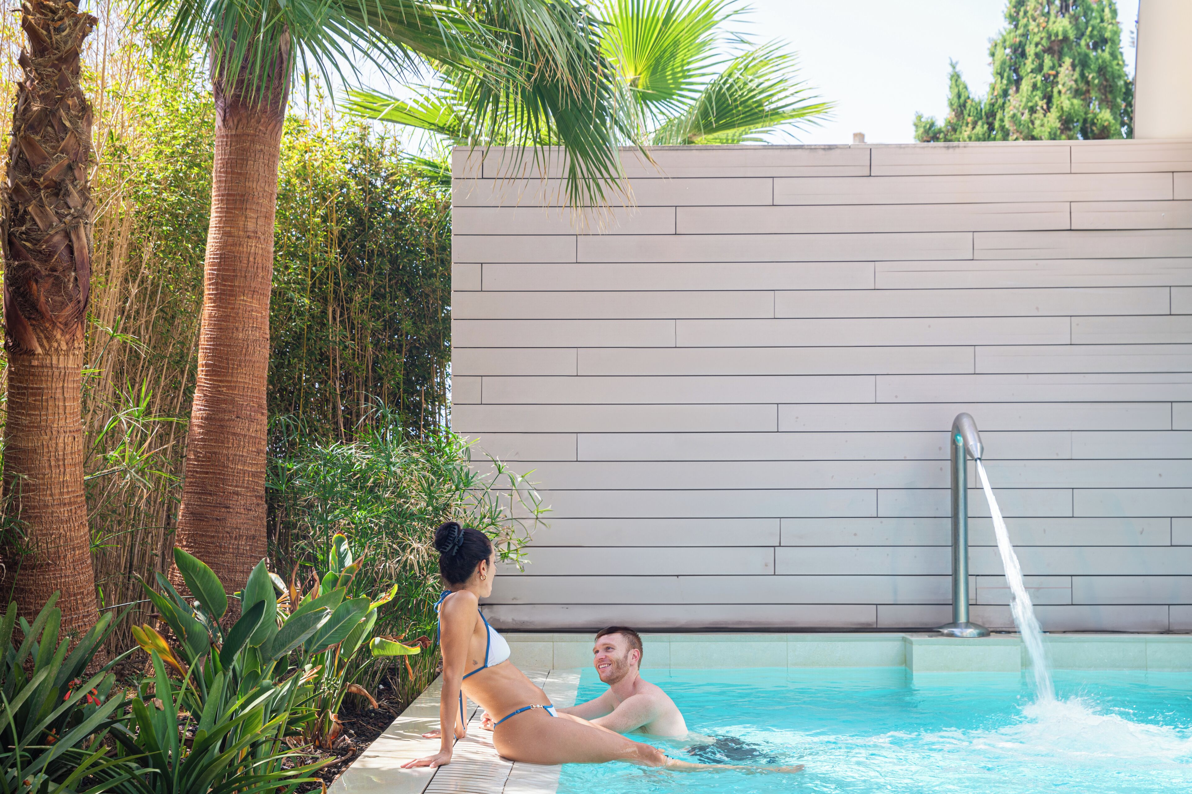 A swimming pool with a water fountain, surrounded by palm trees and greenery.
