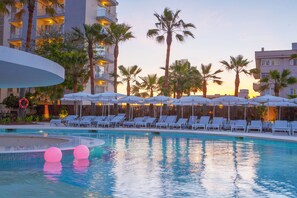 A swimming pool with lounge chairs and umbrellas, surrounded by palm trees and buildings.