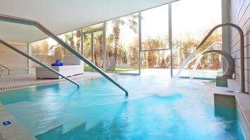 Une piscine intérieure avec des parois en verre transparent, un espace détente blanc et une vue sur les palmiers à l’extérieur.