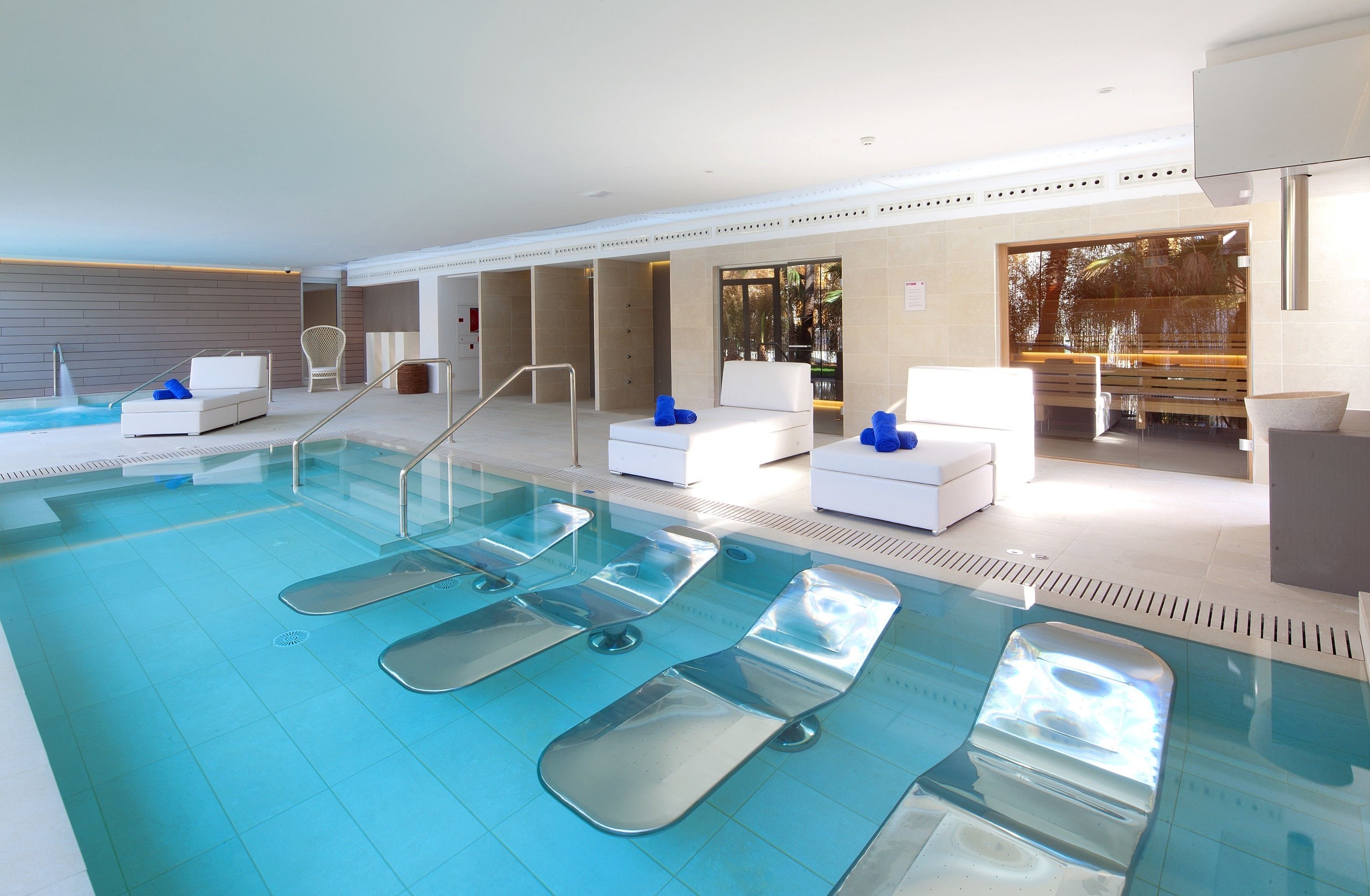 Ein moderner, überdachter Swimmingpool mit klarem Wasser, Liegestühlen und einem durch Glas geschützten Bereich mit Ausblick ins Freie.