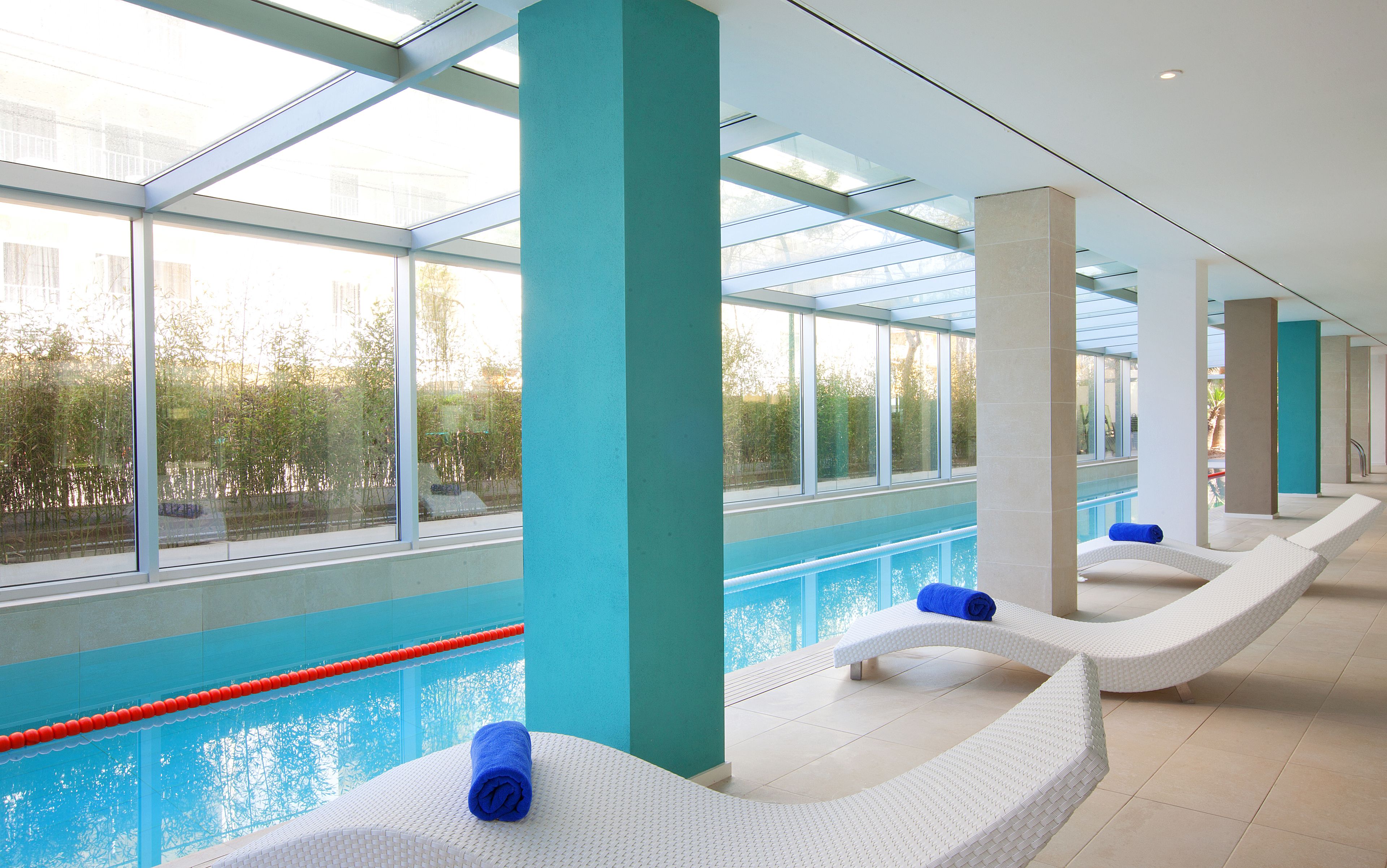 A modern indoor pool area with clear glass walls and ceiling, white loungers with blue towels, and a view of greenery outside.