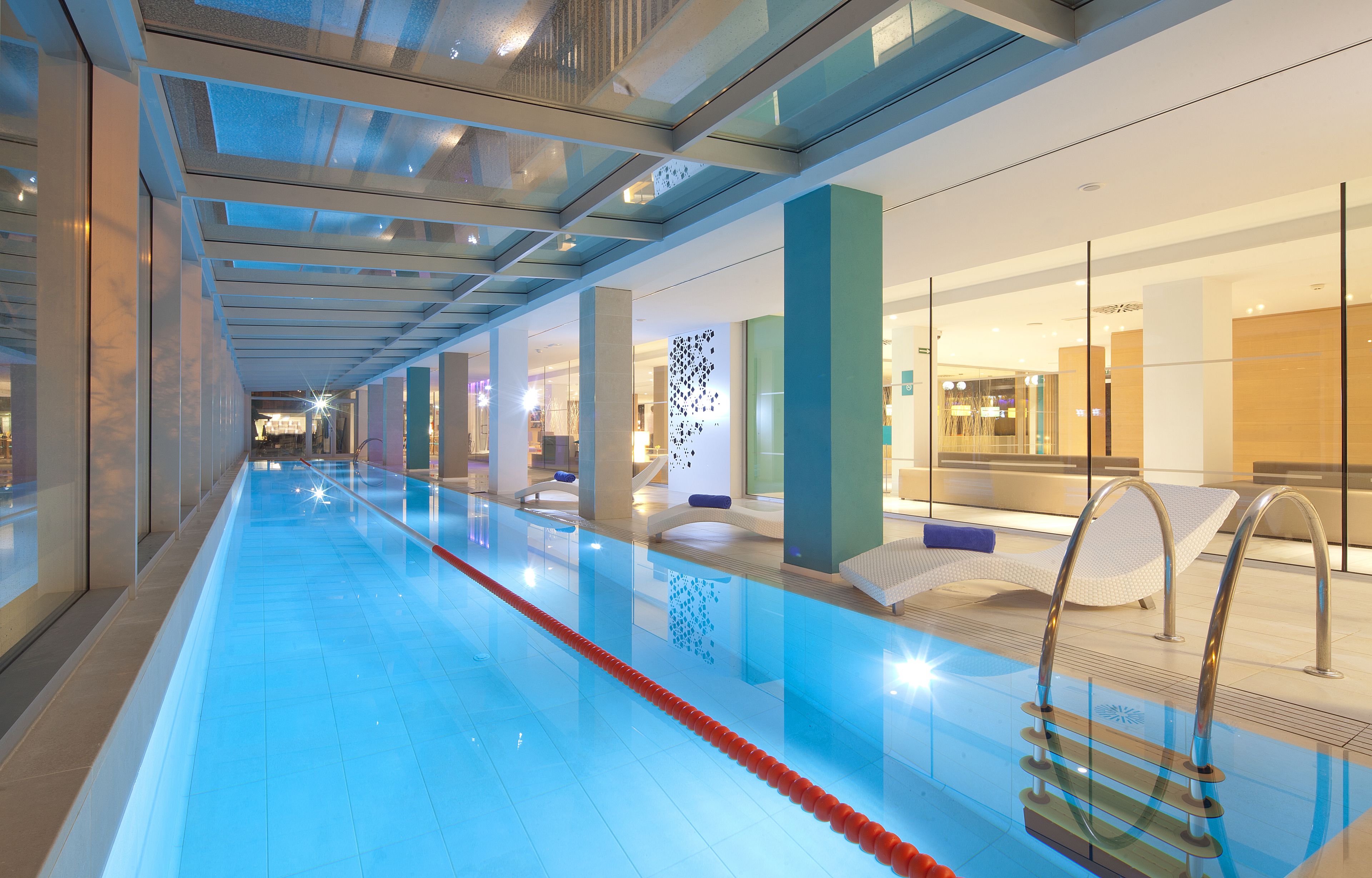 Una larga piscina interior con agua cristalina, rodeada de arquitectura moderna y paredes de vidrio.