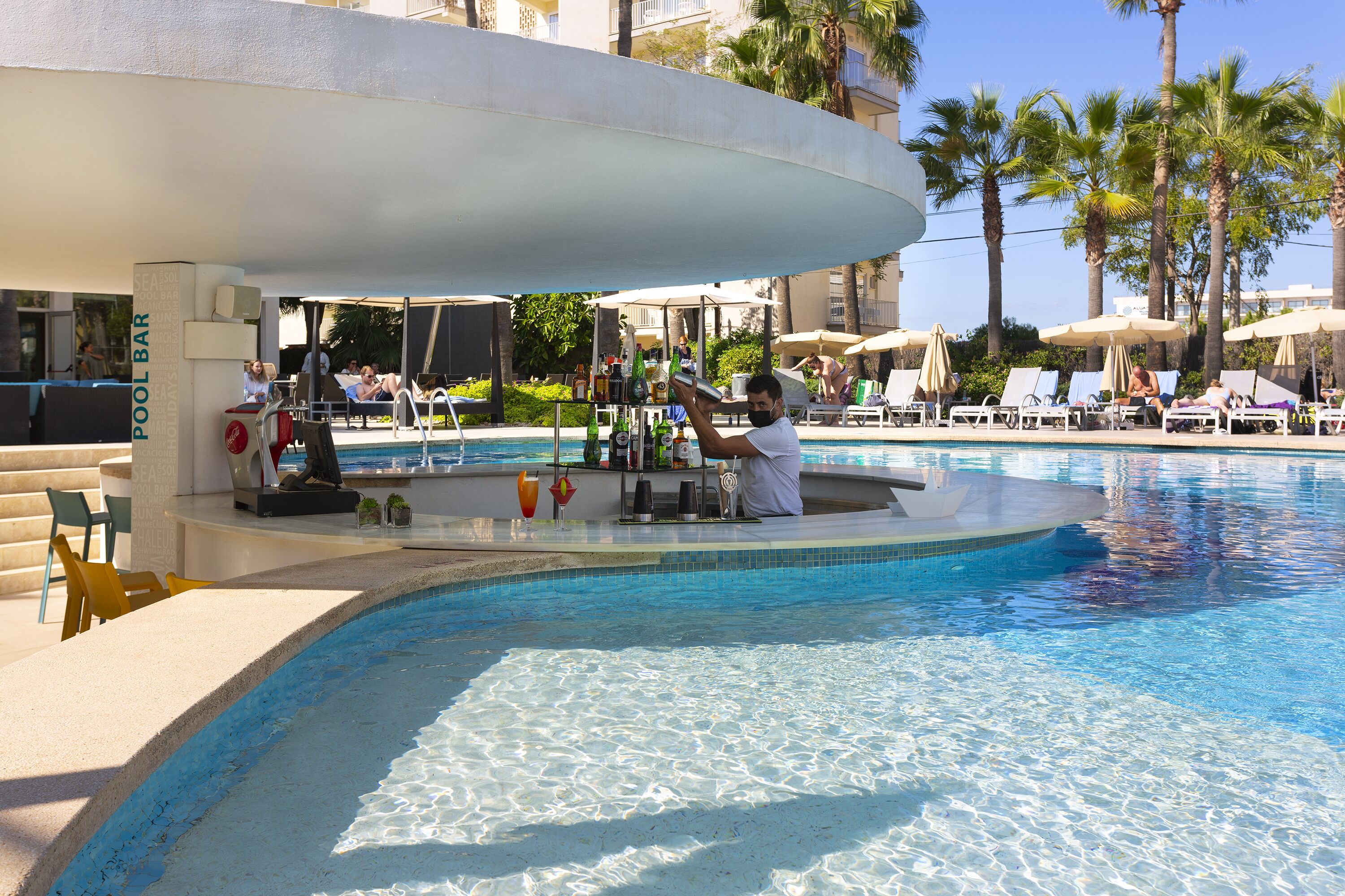 Zona de bar junto a la piscina con sillones de descanso y sombrillas, piscina y palmeras al fondo.