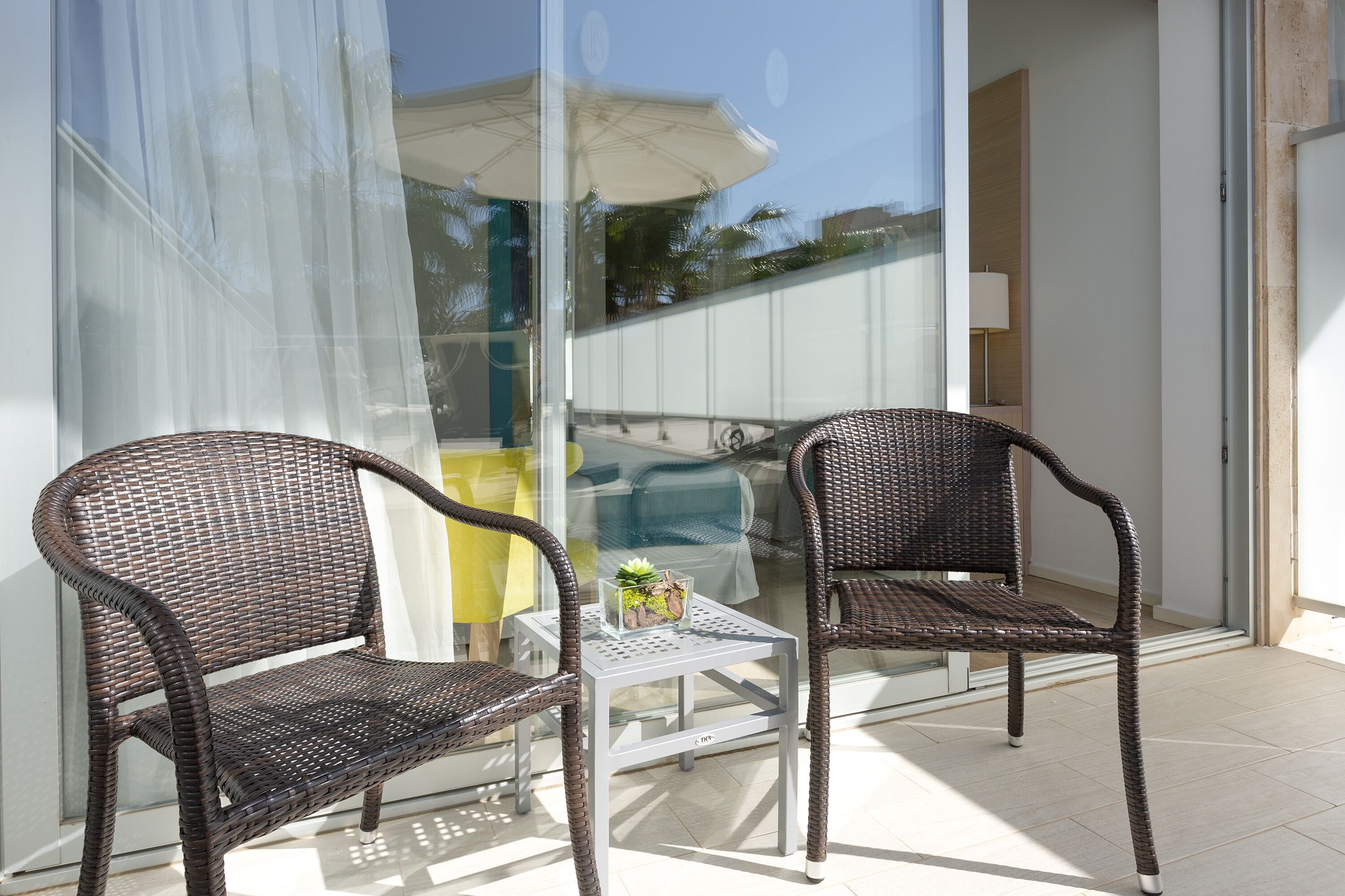 Dos sillas de mimbre y una mesita con superficie de vidrio en un balcón con puerta de cristal y vistas al exterior.