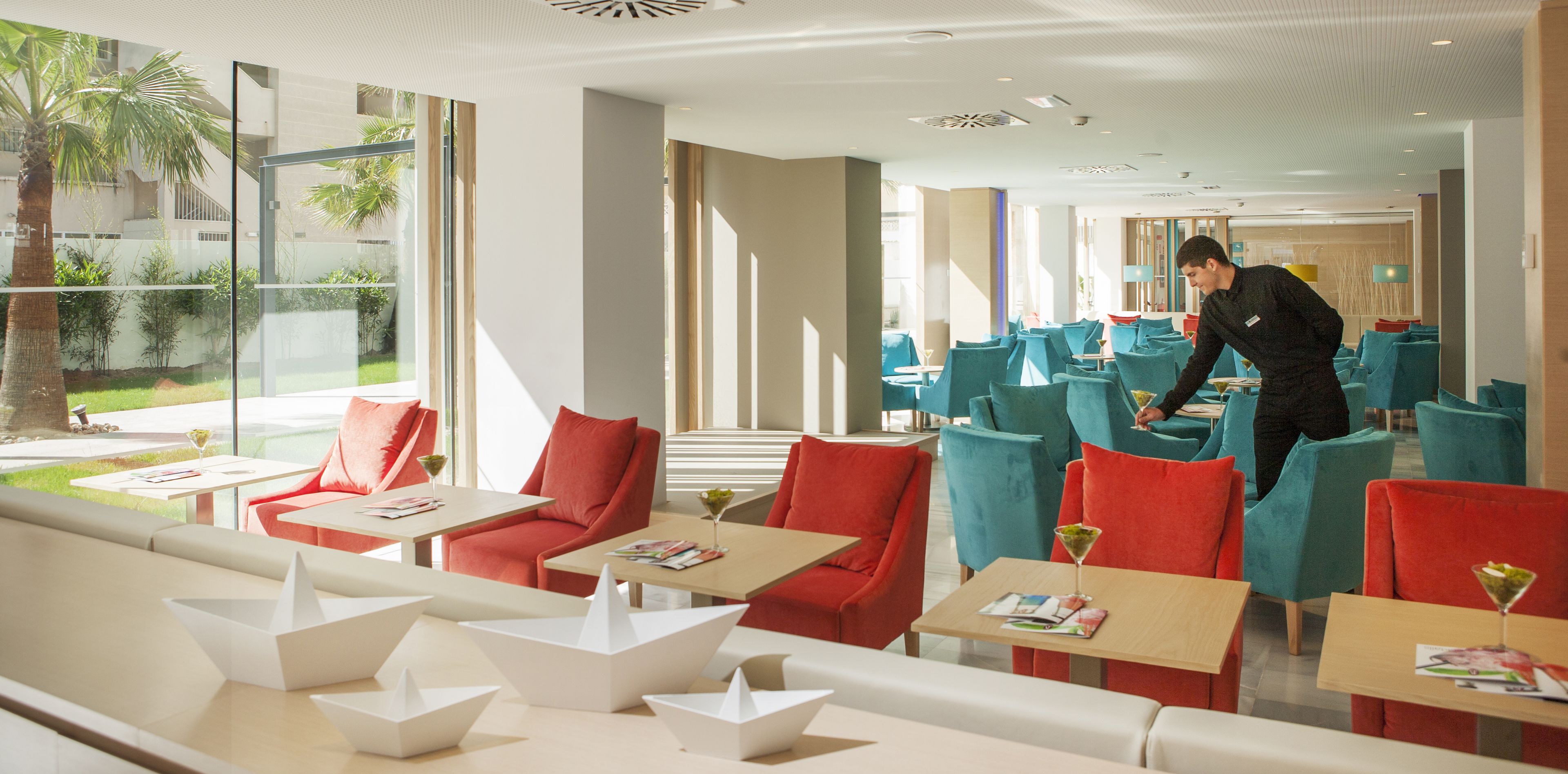 Een moderne hotel lobby met rode en teal zitplaatsen, grote ramen en een ober die gasten bedient.