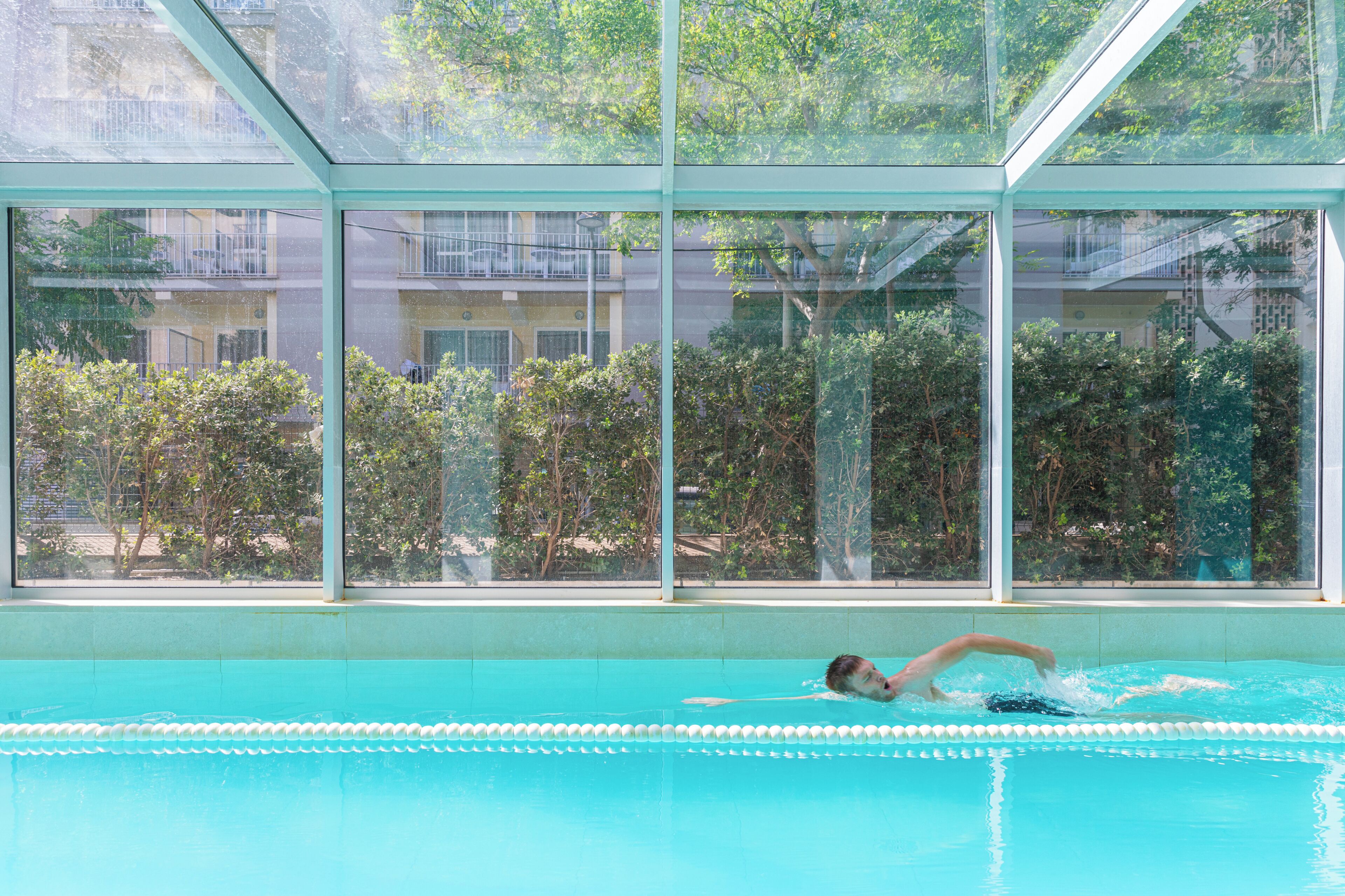 A swimming pool with clear glass walls, a view of greenery, and a building in the background.