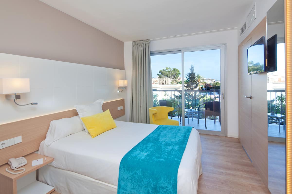 A hotel room with a bed, bedside lamps, a telephone, a balcony with chairs, and a TV mounted on the wall.