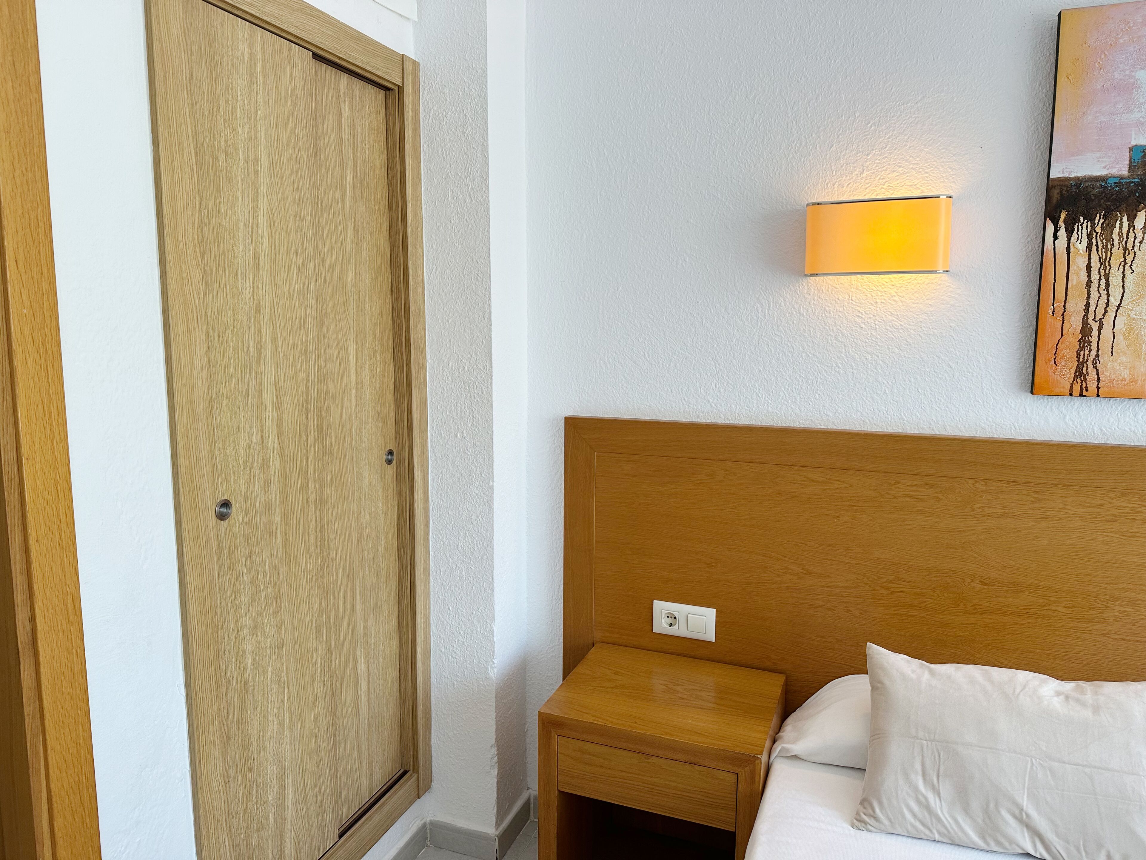 Habitación de hotel con puerta de madera, mesita de noche con lámpara, cama con ropa de cama blanca y un cuadro en la pared.