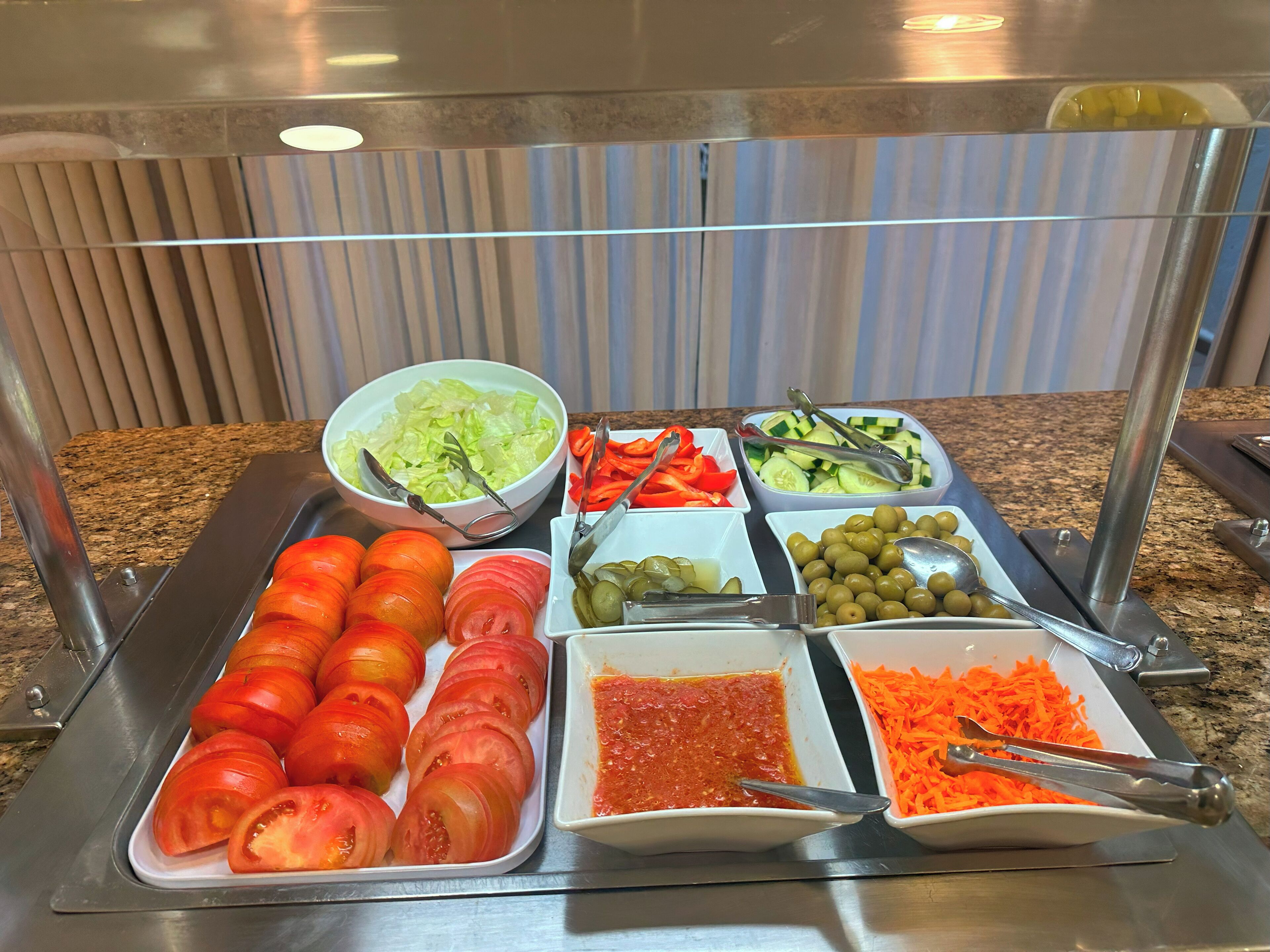 A buffet with various food items including sliced tomatoes, cucumbers, olives, and a bowl of salad.