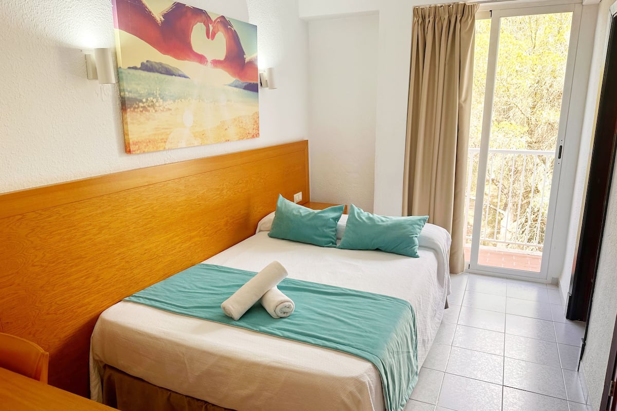 A hotel room with a wooden headboard, a bed with teal bedding, and a painting of hands forming a heart on the wall.