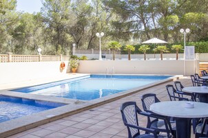 Una piscina con un borde de baldosas, rodeada de sillas y mesas, y palmeras al fondo.