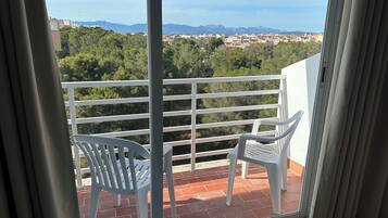 Un balcon avec des chaises blanches et une table, d’où l’on peut admirer un paysage urbain avec des arbres et des immeubles.