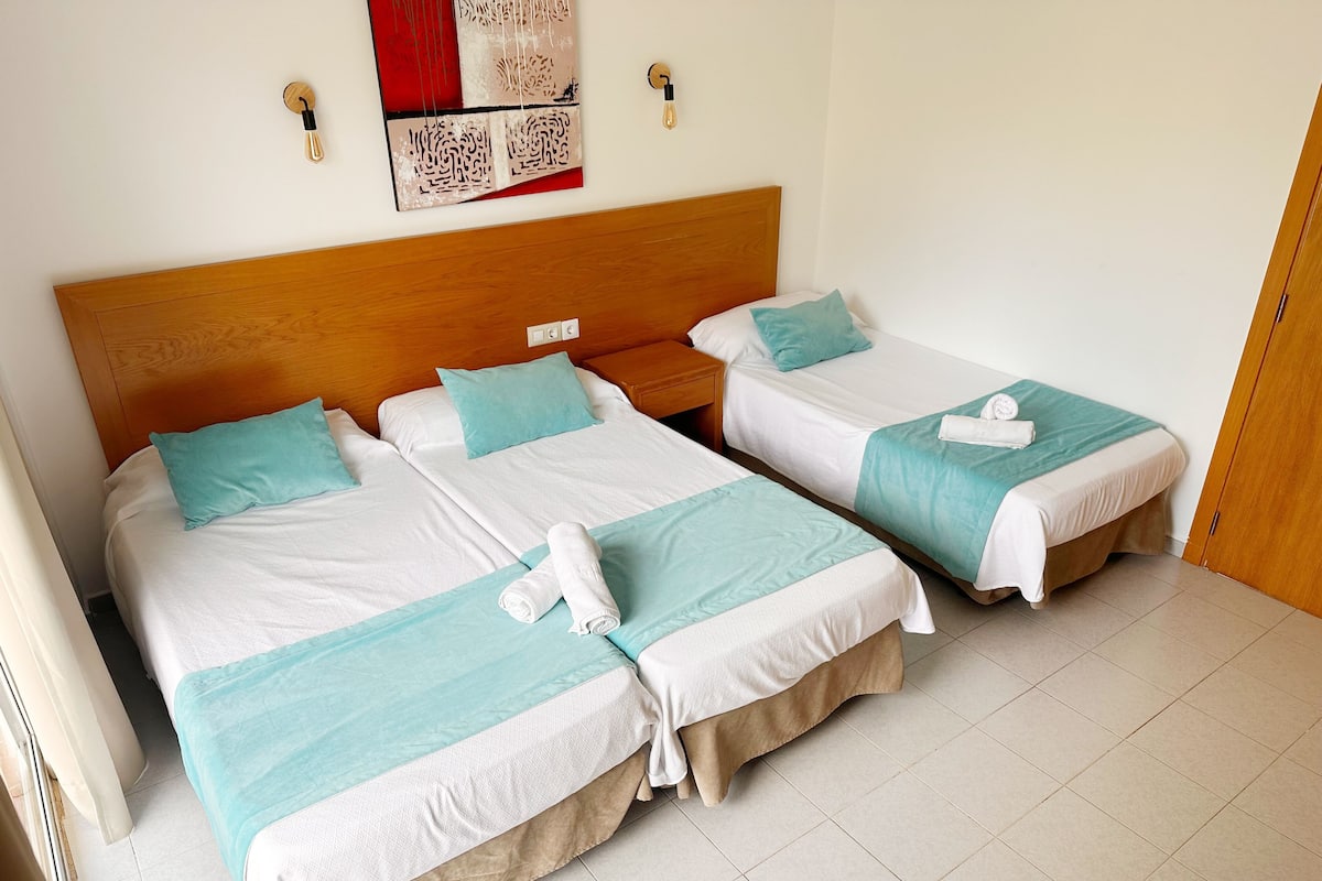 A hotel room with two beds, each with a wooden headboard, white bedding, and teal accents. There is a wall art piece above the beds and a wooden door on the right.