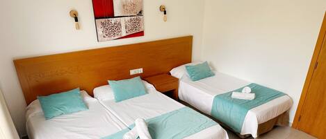 Ein Hotelzimmer mit zwei Betten, jeweils mit einem hölzernen Kopfteil, weißer Bettwäsche und türkisfarbenen Akzenten. Darüber hängen Bilder an der Wand und rechts ist eine Holztür zu sehen.