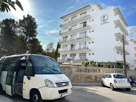 Una camioneta blanca con la marca 'Hotel Selva Arena' estacionada frente a un edificio de varios pisos con balcones y cielo despejado.