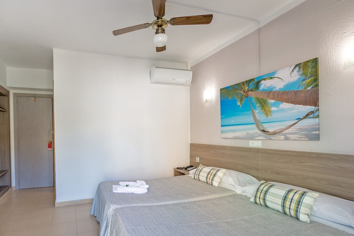 Ein Hotelzimmer mit einem Bett, einem Deckenventilator, einer Klimaanlage, einem Bild mit Strandmotiv und einer Tür.