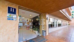 Entrada de un hotel con puerta de vidrio transparente, un letrero con una 'H' grande y un felpudo que dice 'Hotel Costa Mediterranea'.