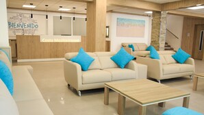 Un moderno hall de hotel con sofás color beige, cojines azules y un mostrador de recepción de madera.