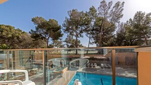 Zona de piscina con paredes de vidrio transparente, sillas de descanso y vistas a árboles y edificios.