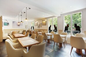 A modern restaurant with white walls, beige chairs, and tables. There are large windows with a view of trees. - Hôtel Le Chat Noir (Paris)