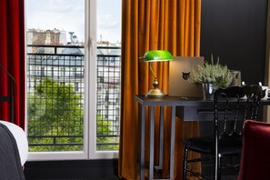 A room with a view of buildings and greenery through a window, a desk with a lamp, a chair, and a potted plant. - Hôtel Le Chat Noir (Paris)
