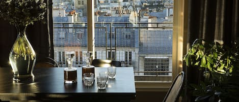 Une table à manger avec un vase et des verres, et une vue sur le paysage urbain à travers une fenêtre ornée d’une rambarde en métal.