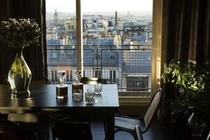 Une table à manger avec un vase et des verres, et une vue sur le paysage urbain à travers une fenêtre ornée d’une rambarde en métal.
