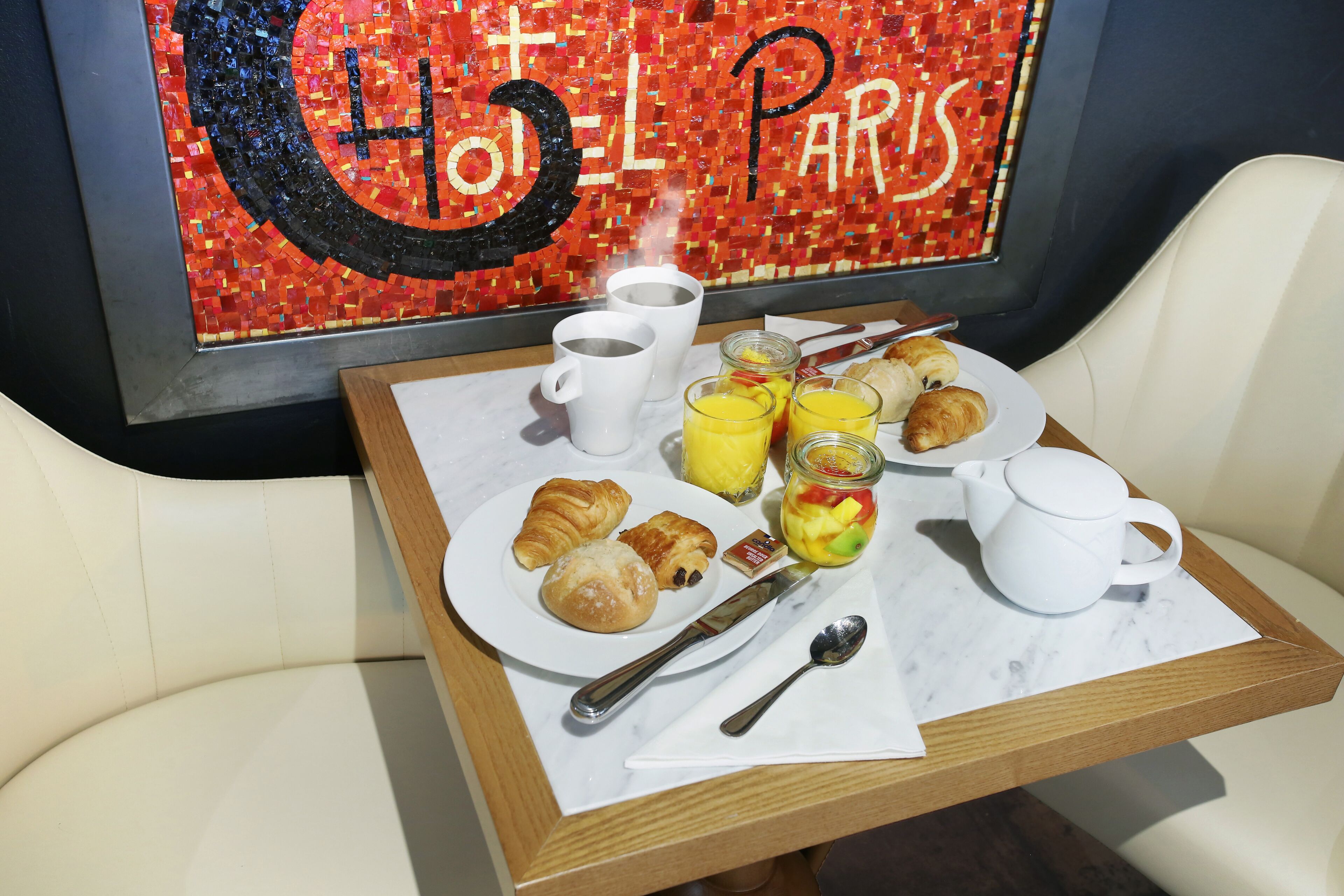 Un vassoio per la colazione con croissant, succo di frutta e una tazza di caffè su un tavolo, di fronte a una parete con un cartello in mosaico che riporta la scritta 'Hotel Paris'.