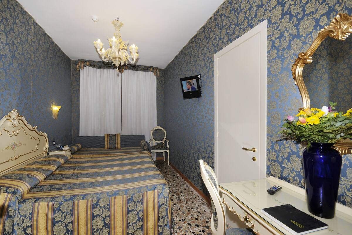 Une chambre d’hôtel avec un lit, un bureau, une chaise, un miroir, un vase de fleurs et un lustre.